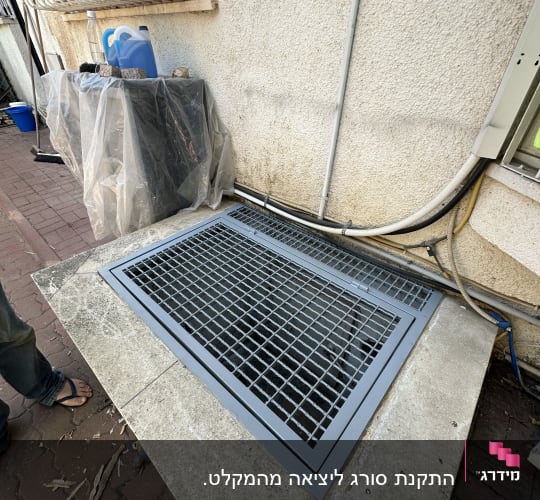 סורג מתכת מותקן על פתח ברצפה ליד קיר חיצוני